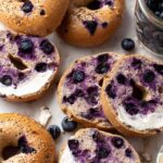 Top view of several sliced and whole gluten free blueberry bagels on a white background with scattered fresh blueberries.