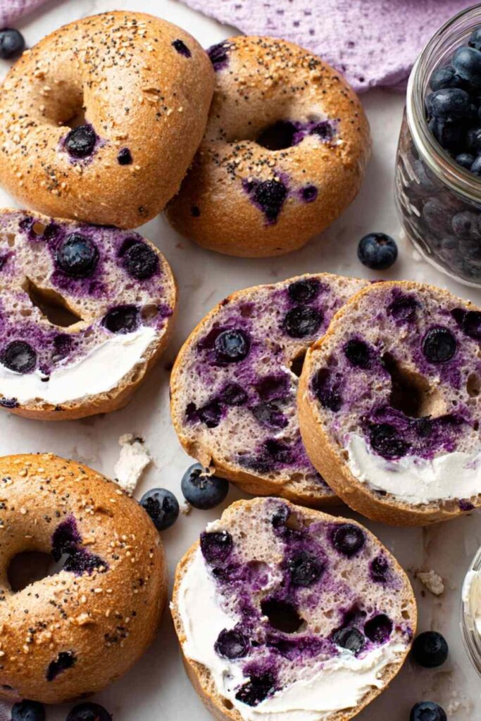 Top view of several sliced and whole gluten free blueberry bagels on a white background with scattered fresh blueberries.