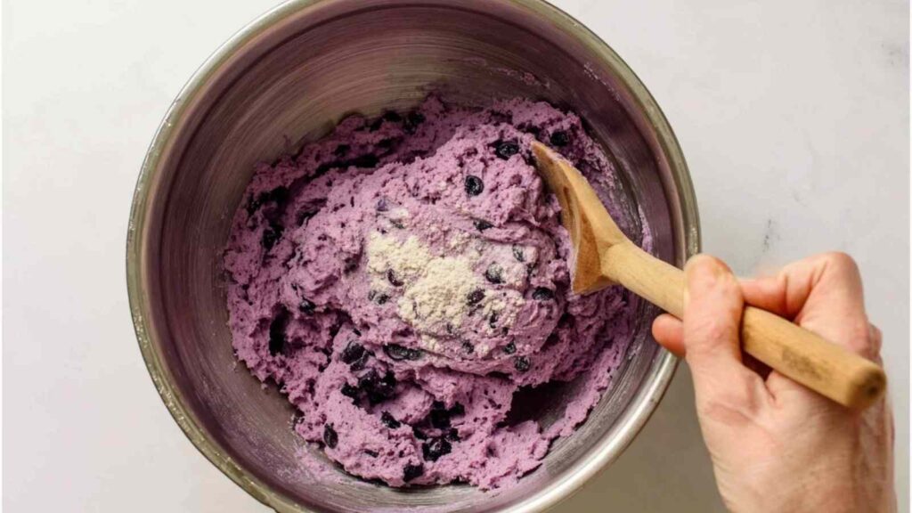 Hand mixing fresh blueberries into purple-hued gluten free bagel dough in a metal bowl.