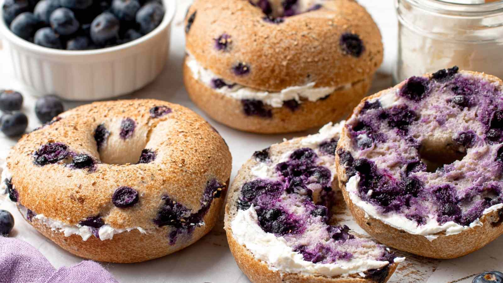 Golden brown gluten free blueberry bagels filled with fresh blueberries and slathered with cream cheese, styled with a bowl of blueberries on the side.