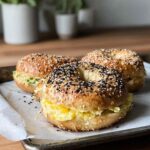 Three Greek yogurt bagel breakfast sandwiches filled with scrambled eggs and avocado on a tray.