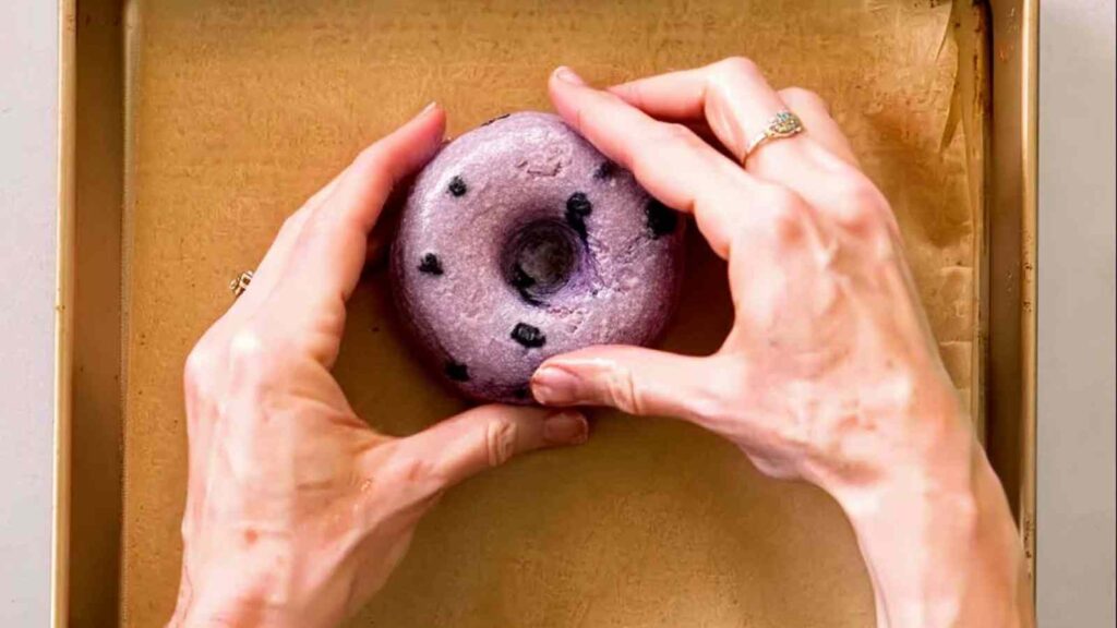 Hands shaping a round gluten free blueberry bagel dough ball with a center hole on a parchment-lined tray.