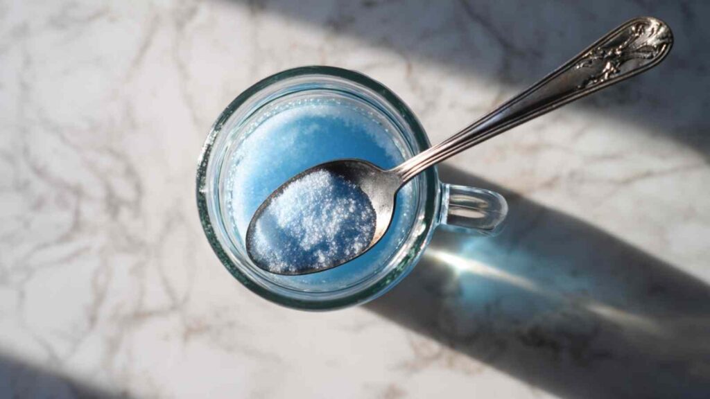 blue salt energy drink 9 A silver spoon holding a heap of fine white powder, such as Persian blue salt or ashwagandha powder, positioned over a glass mug containing a light blue liquid, a step in preparing the "salt trick for men in bed recipe.