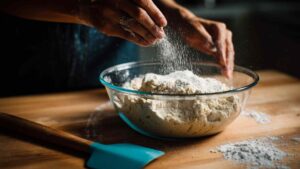 Hands sprinkling flour into a glass bowl of sourdough dough during the early mixing stage
