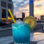 A refreshing blue cocktail with ice, a lemon wheel garnish, and a yellow straw in a textured glass, set against a blurred city skyline at dusk with a prominent blue-lit building, showcasing the final presentation of the "salt trick for men in bed recipe" as a pre-bedtime beverage.