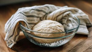 Risen bolted flour sourdough dough in a glass bowl, lightly dusted with flour and covered by a striped kitchen towel