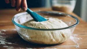 Bolted flour sourdough dough rising in a glass bowl with a blue spatula resting inside
