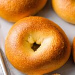 Close-up of shiny, golden egg bagels on a tray, one sliced and spread with cream cheese