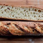 Whole sourdough baguette with caramelized crust and a cross-section showing open, airy crumb on a wooden cutting board