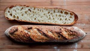Whole sourdough baguette with caramelized crust and a cross-section showing open, airy crumb on a wooden cutting board