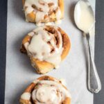 Three glazed sourdough cinnamon rolls on parchment with a spoon of cream cheese icing