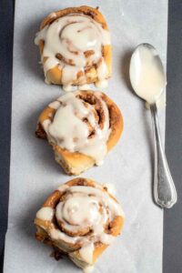 Three glazed sourdough cinnamon rolls on parchment with a spoon of cream cheese icing