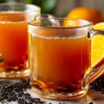 Two clear mugs filled with golden Costa Rican tea surrounded by fresh oranges and tea leaves on a marble counter.