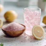 Glass of pink detox drink with fresh lemon halves, pink salt in a wooden bowl, and scattered salt crystals on a light kitchen surface