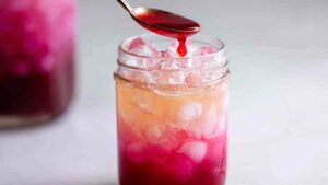 Close-up of red syrup being poured into a mason jar of ice and soda, showing a beautiful color gradient forming.