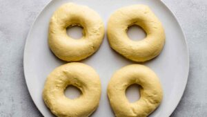 Four raw onion bagel dough rings arranged on a plate, ready for the boiling step