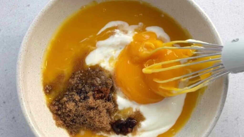 Mixing bowl filled with pumpkin puree, yogurt, eggs, and brown sugar being whisked together