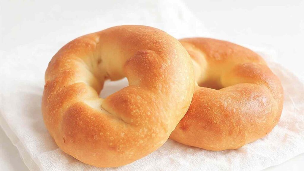 Two plain, untoasted bagels on a white linen, illustrating the starting state before slicing and baking.