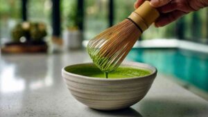 Close-up of bamboo whisk frothing matcha in a ceramic bowl on a white kitchen counter.