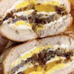 Close-up of a bagel sandwich cut in half, showcasing the rich, dark homemade bacon jam, a bright yellow fried egg yolk, and a thick layer of white cheese.