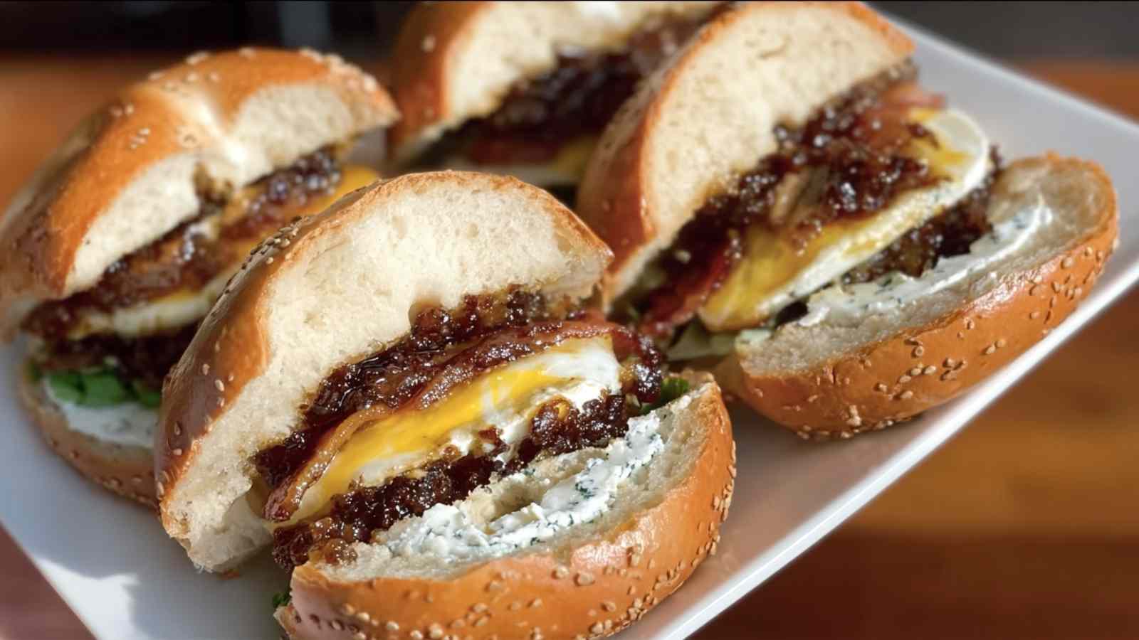 Four bagel sandwiches on a white plate, each cut in half to display layers of homemade bacon jam, a fried egg with a visible yolk, creamy white cheese, and fresh green lettuce on a sesame seed bagel.