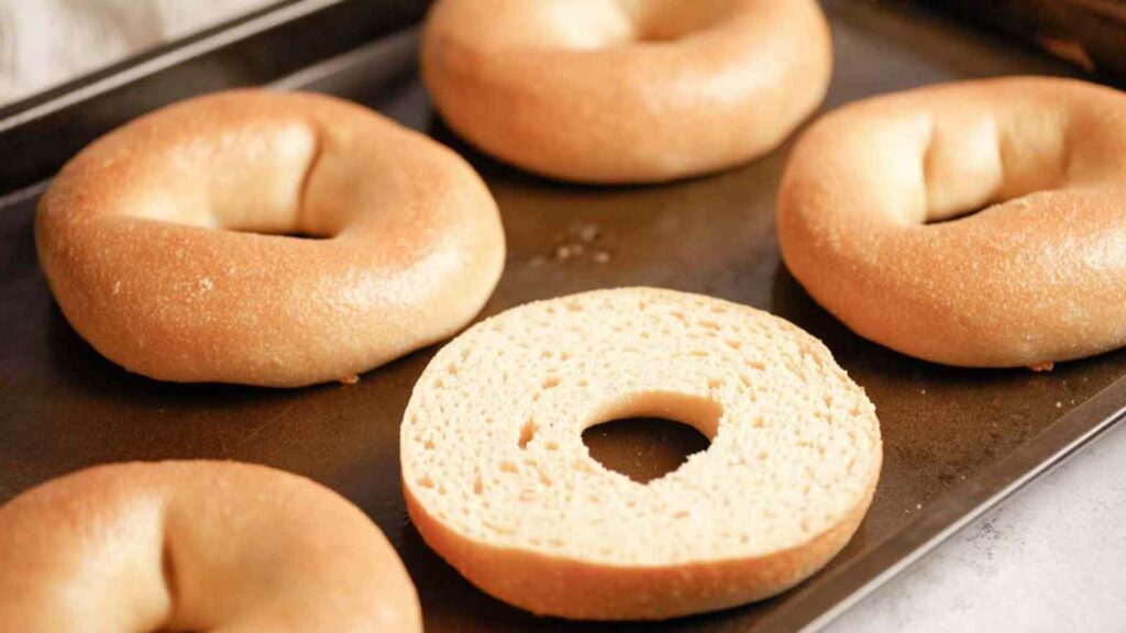 A baking sheet with several whole bagels and one sliced bagel half, ready for oven toasting.