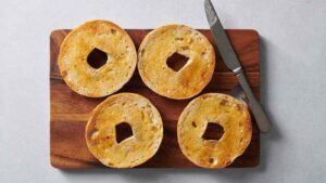 Four golden-brown oven-toasted bagel halves on a wooden cutting board with a knife, showcasing crispy texture.