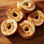 Five oven-toasted bagel halves, some with seasoning, arranged on a wooden board, highlighting the golden-brown finish.