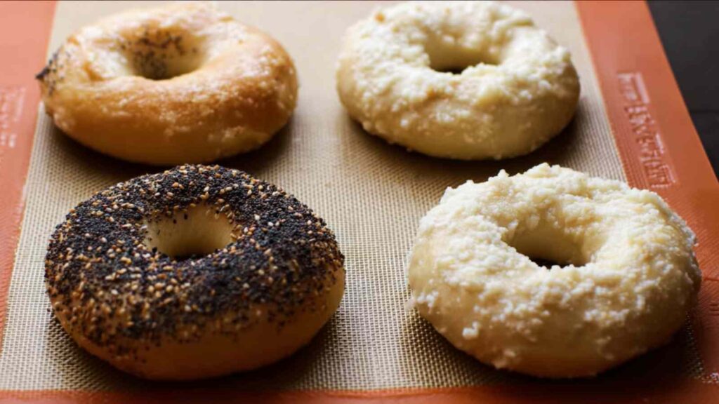 Four golden-brown baked bagels with poppy seeds, sesame seeds, and plain toppings on a baking mat.