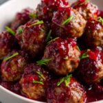 Bowl of cranberry-glazed beef meatballs topped with rosemary, set against glowing holiday lights—ideal appetizer for Christmas gatherings.