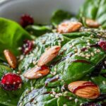 Close-up of spinach leaves topped with cranberries, almonds, sesame seeds, and balsamic glaze in a white bowl—vibrant and festive holiday salad.