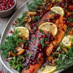 Salmon fillet topped with pomegranate glaze and seeds, garnished with fresh dill and lemon slices, served on a silver holiday platter.
