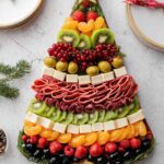 Charcuterie board in shape of a Christmas tree made with stacked layers of cheddar, grapes, salami, olives, mozzarella, and herbs on white background