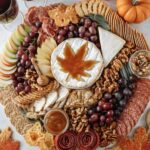 Vertical view of fall charcuterie board with maple leaf brie, wine glasses, grapes, apples, crackers, cured meats, and mini pumpkins