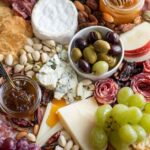 Vertical close-up of a colorful charcuterie board with green and red grapes, Brie cheese, marinated olives, honey jar, and mixed nuts