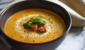 Creamy roasted veggie soup in white bowl topped with fresh basil and black pepper