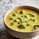 Creamy broccoli cheddar soup in a white bowl topped with croutons and shredded cheese.