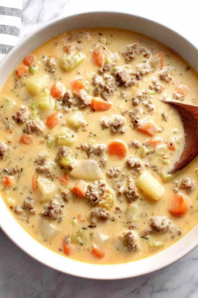 Cheeseburger soup in a white bowl topped with shredded cheese, pickles, and diced tomatoes