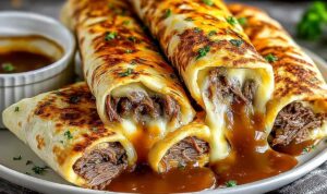 French dip tortilla roll-ups sliced diagonally on a wooden board with a small bowl of au jus dipping sauce