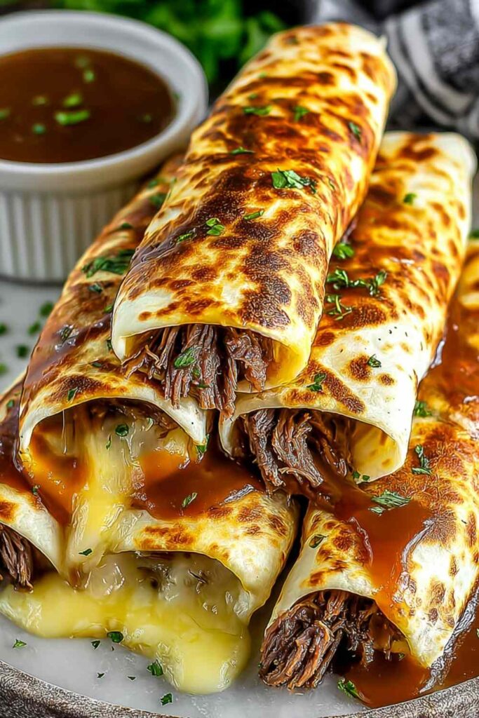 French dip tortilla roll-ups sliced diagonally on a wooden board with a small bowl of au jus dipping sauce
