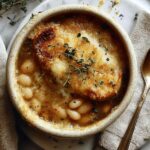 A bowl of French Onion and Butter Bean Soup topped with melted Gruyere cheese and toasted baguette slices