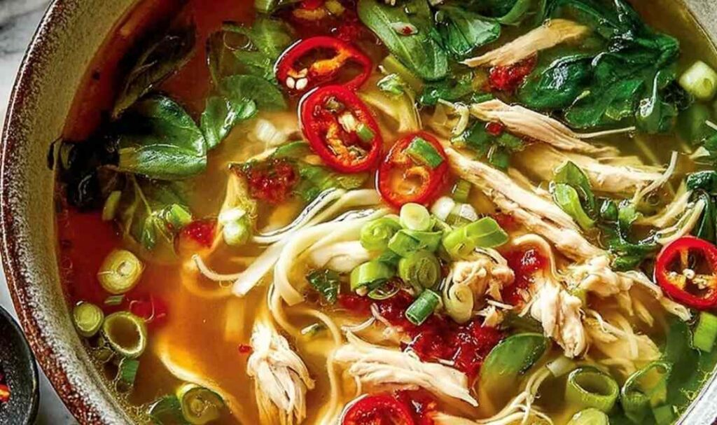 Bowl of ginger garlic chicken noodle soup with egg noodles, julienned carrots, wilted spinach, and green onion garnish