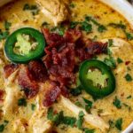 Spicy jalapeno popper chicken soup in a white bowl topped with shredded cheddar cheese, crispy bacon bits, and fresh sliced jalapenos