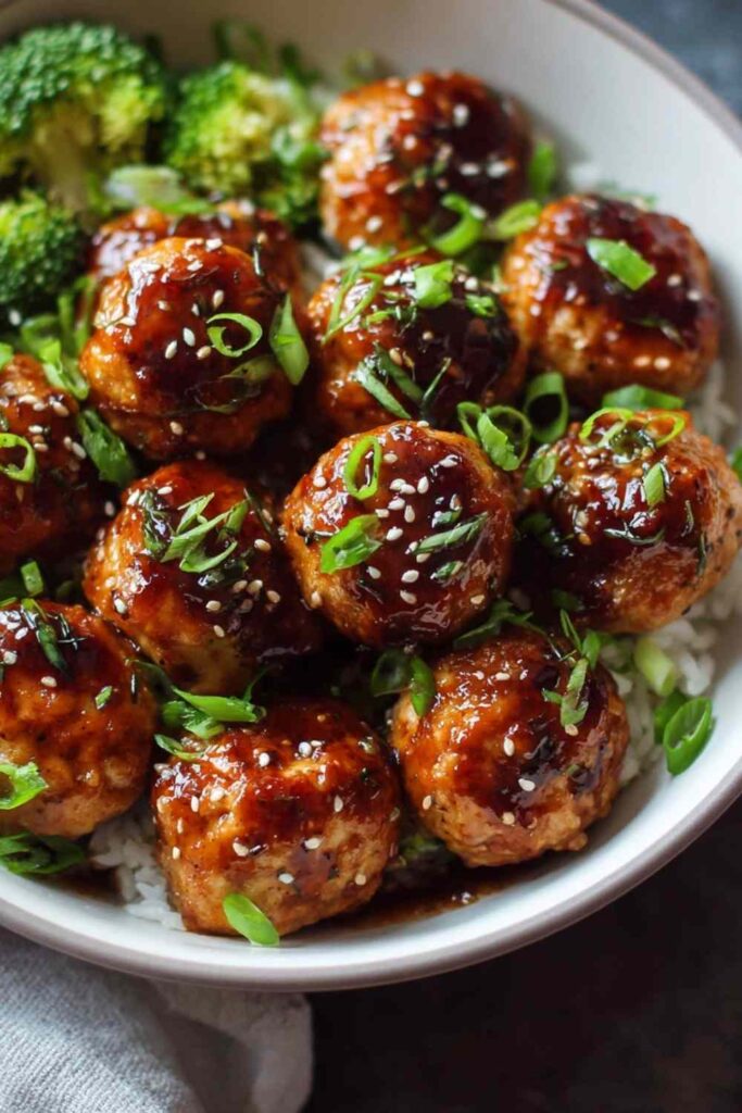 Baked Asian chicken meatballs coated in sticky honey sesame glaze garnished with toasted sesame seeds and green onions in a white bowl over rice