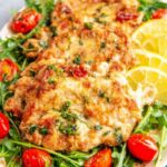 Chicken paillard recipe on a white plate with arugula salad, cherry tomatoes, and lemon wedges