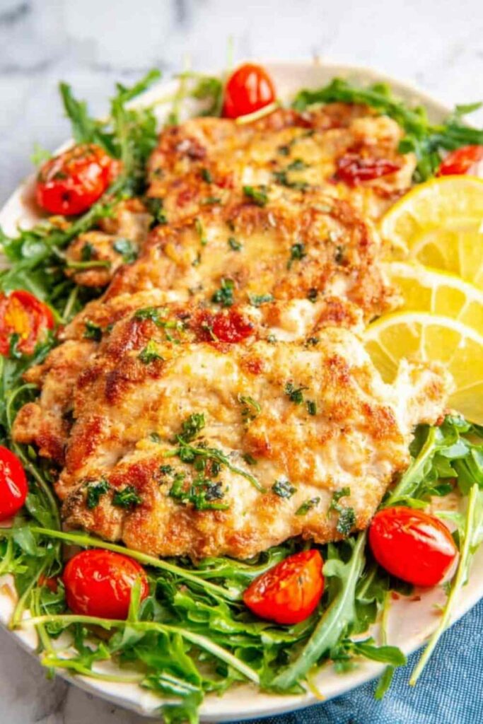Chicken paillard recipe on a white plate with arugula salad, cherry tomatoes, and lemon wedges
