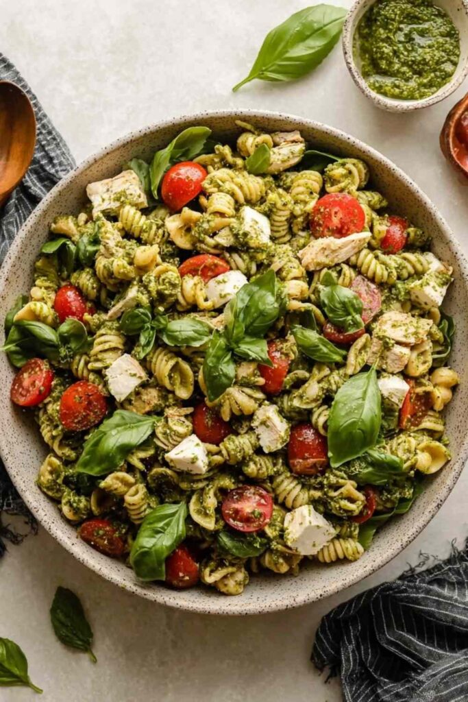 Chicken pesto pasta salad in a large white bowl with cherry tomatoes, cucumber, and fresh basil