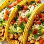 Juicy shredded chicken tacos topped with fresh cilantro, diced tomatoes, shredded cheese, sour cream, and lime wedges on a wooden serving board