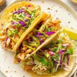 Copycat cantina chicken tacos loaded with shredded chicken, cheddar cheese, lettuce, purple cabbage, pico de gallo, and avocado salsa verde on a wooden serving board