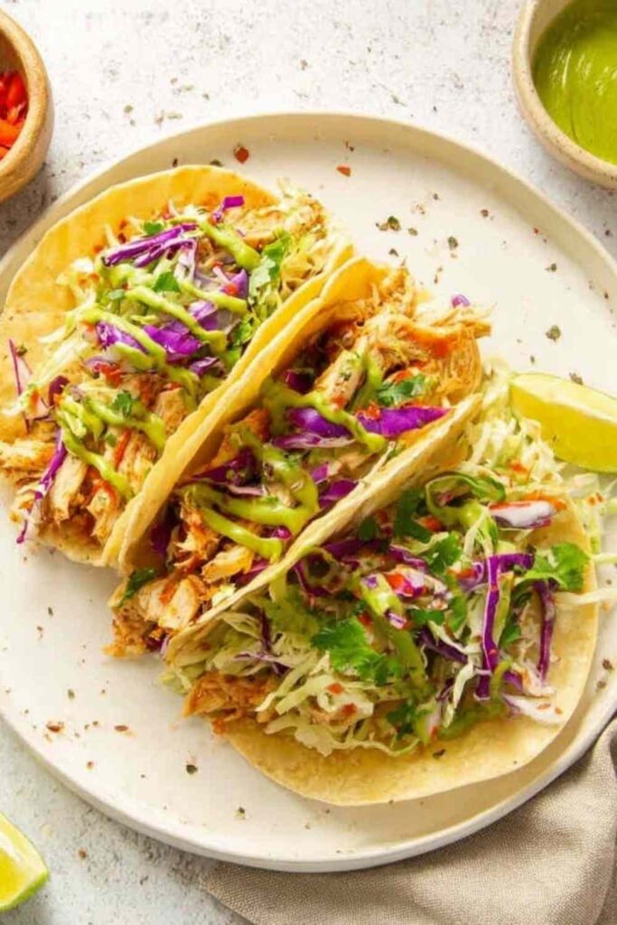 Copycat cantina chicken tacos loaded with shredded chicken, cheddar cheese, lettuce, purple cabbage, pico de gallo, and avocado salsa verde on a wooden serving board